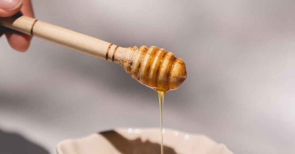 Hand holds a honey dipper dripping honey into a ceramic bowl with leaves around.